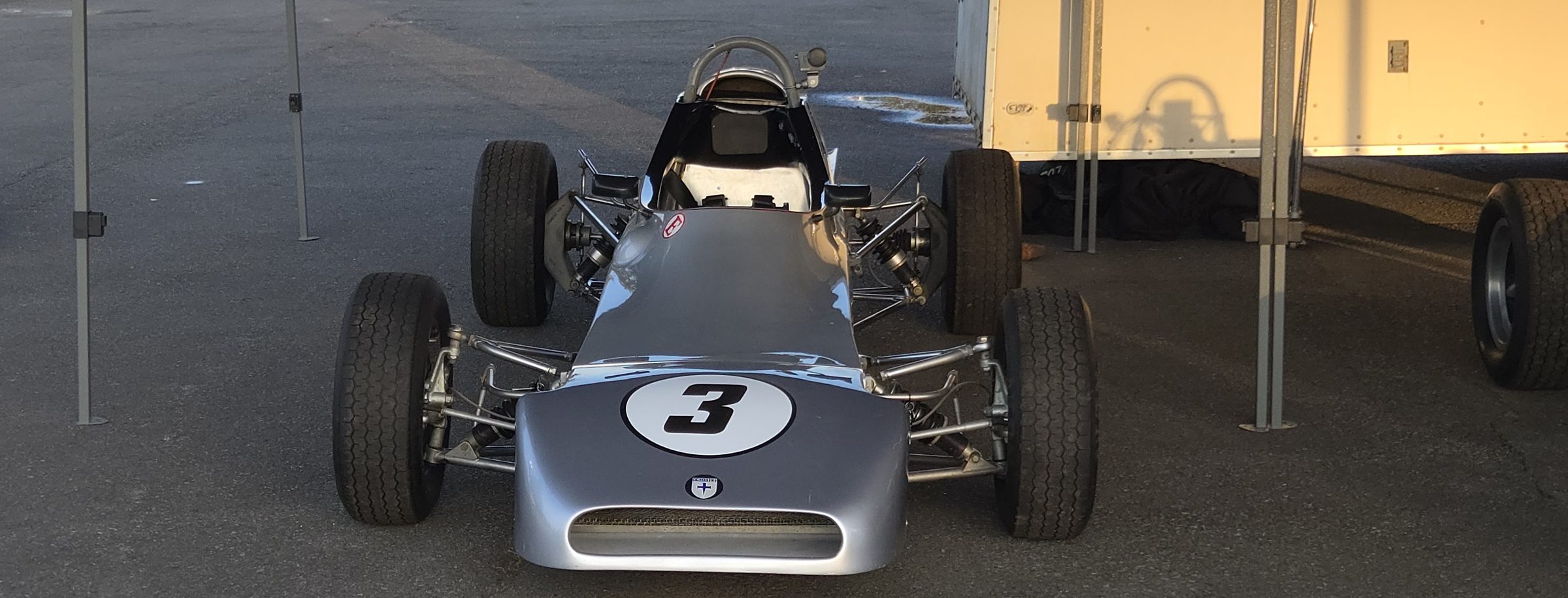 The silver number 3 Crosslé 35F parked head-on under a paddock tent in morning light